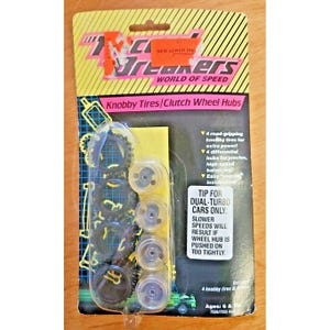 Pode incluir: Um pacote de peças de brinquedo Track Breakers World of Speed contendo quatro pneus pretos com cravos e quatro cubos de roda transparentes. A embalagem apresenta listras diagonais amarelas e pretas, texto rosa e instruções.