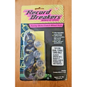 Pode incluir: Um conjunto de acessórios de brinquedo Record Breakers World of Speed num suporte de cartão. A embalagem contém quatro pneus de corrida pretos e quatro cubos de roda transparentes. O texto na embalagem inclui características como pneus de corrida que guincham e uma dica para carros de turbo duplo sobre a instalação. O design apresenta um padrão de grelha amarelo e preto.