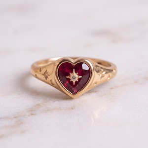 Könnte beinhalten: Ein goldener Ring mit einem herzförmigen, dunkelroten Edelstein in der Mitte. Ein kleiner, klarer, rund geschliffener Stein ist in die Mitte des roten Herzens eingelassen. Der Goldring ist mit kleinen Stern- und Mondsichelmotiven graviert.