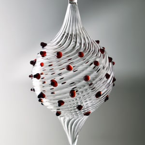 May include: Clear glass ornament with a spiral design and red accents. The teardrop-shaped ornament has a red loop at the top for hanging and is decorated with small, red, rounded shapes. A festive, decorative item.