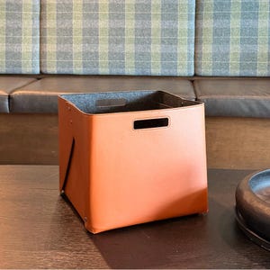 May include: A square storage bin featuring a smooth, tan-colored exterior and a dark gray felt interior lining. The bin includes integrated rectangular cut-out handles on the front and back panels, with visible metal rivets securing the side folds.
