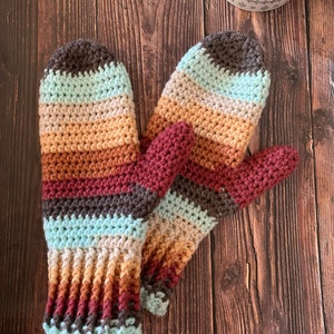 May include: A pair of striped crocheted mittens in shades of brown, beige, red, blue, and gray. The mittens are laid out on a wooden surface.