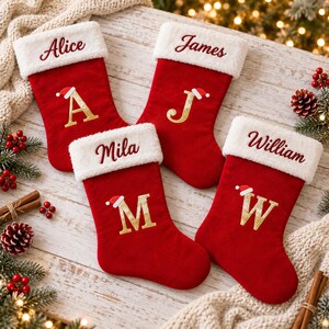 May include: Four red Christmas stockings with white faux fur cuffs. Each stocking features a name embroidered in red script and a large gold letter monogram topped with a small Santa hat. The names are Alice, James, Mila, and William.