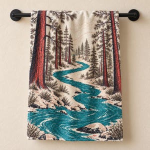 May include: A rectangular towel featuring a printed illustration of a winding teal river flowing through a forest of tall red-barked pine trees. The scene includes rocky banks and evergreen trees set against a cream-colored background.