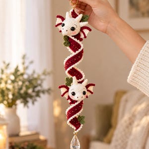 May include: A crochet hanging ornament featuring two white dragon heads with black eyes and pink cheeks, connected by a long, spiraling dark red ribbon with white edges, accented with small green leaves and a clear teardrop crystal at the bottom.
