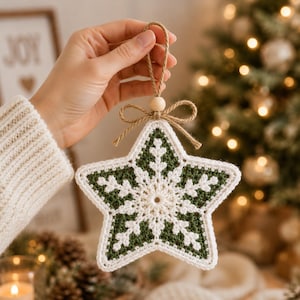 May include: A star-shaped crochet ornament featuring a dark green background with a white snowflake pattern. The ornament is topped with a small wooden bead and a jute twine bow, with a matching jute loop for hanging from a Christmas tree.