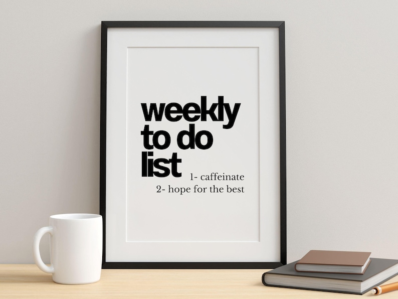May include: A framed print with the words "weekly to do list" in bold black font. The list includes "1- caffeinate" and "2- hope for the best." A white mug, a closed book, and a pencil are on the table.