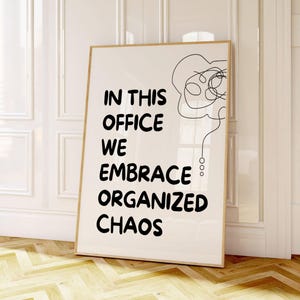 Op de afbeelding: Een ingelijste print met de tekst "IN THIS OFFICE WE EMBRACE ORGANIZED CHAOS" in het zwart. De print heeft een minimalistisch ontwerp met een lijntekening en wordt gepresenteerd tegen een witte muur en een houten vloer.