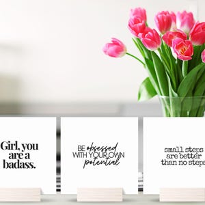 May include: Three white cards with inspirational quotes printed in black. The quotes are "Girl, you are a badass.", "Be obsessed with your own potential", and "Small steps are better than no steps".