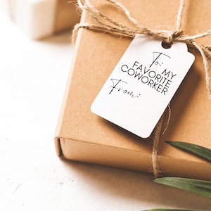 Pu&ograve; includere: Una scatola regalo marrone con un'etichetta regalo bianca che dice "A: Il mio collega preferito Da: ". La scatola &egrave; legata con lo spago e ha una foglia verde in cima.