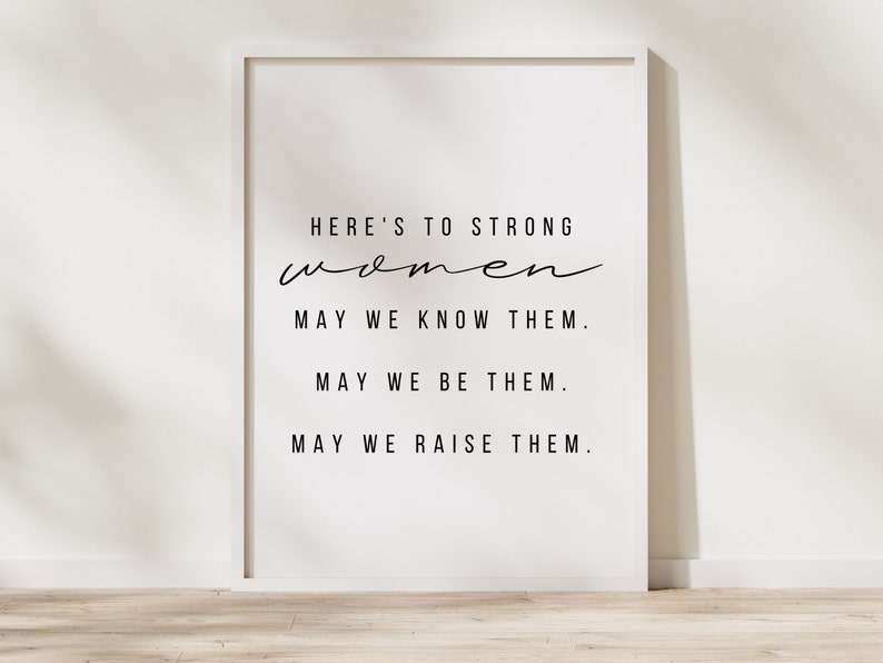 May include: A white framed print with black text that reads "HERE'S TO STRONG women MAY WE KNOW THEM. MAY WE BE THEM. MAY WE RAISE THEM."