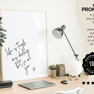 May include: A framed print with a white background and black text that reads "Life is tough my darling, but so are you." The print is on a desk with a laptop, a lamp, and a potted plant.