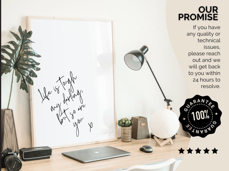 May include: A framed print with a white background and black text that reads "Life is tough my darling, but so are you." The print is on a desk with a laptop, a lamp, and a potted plant.