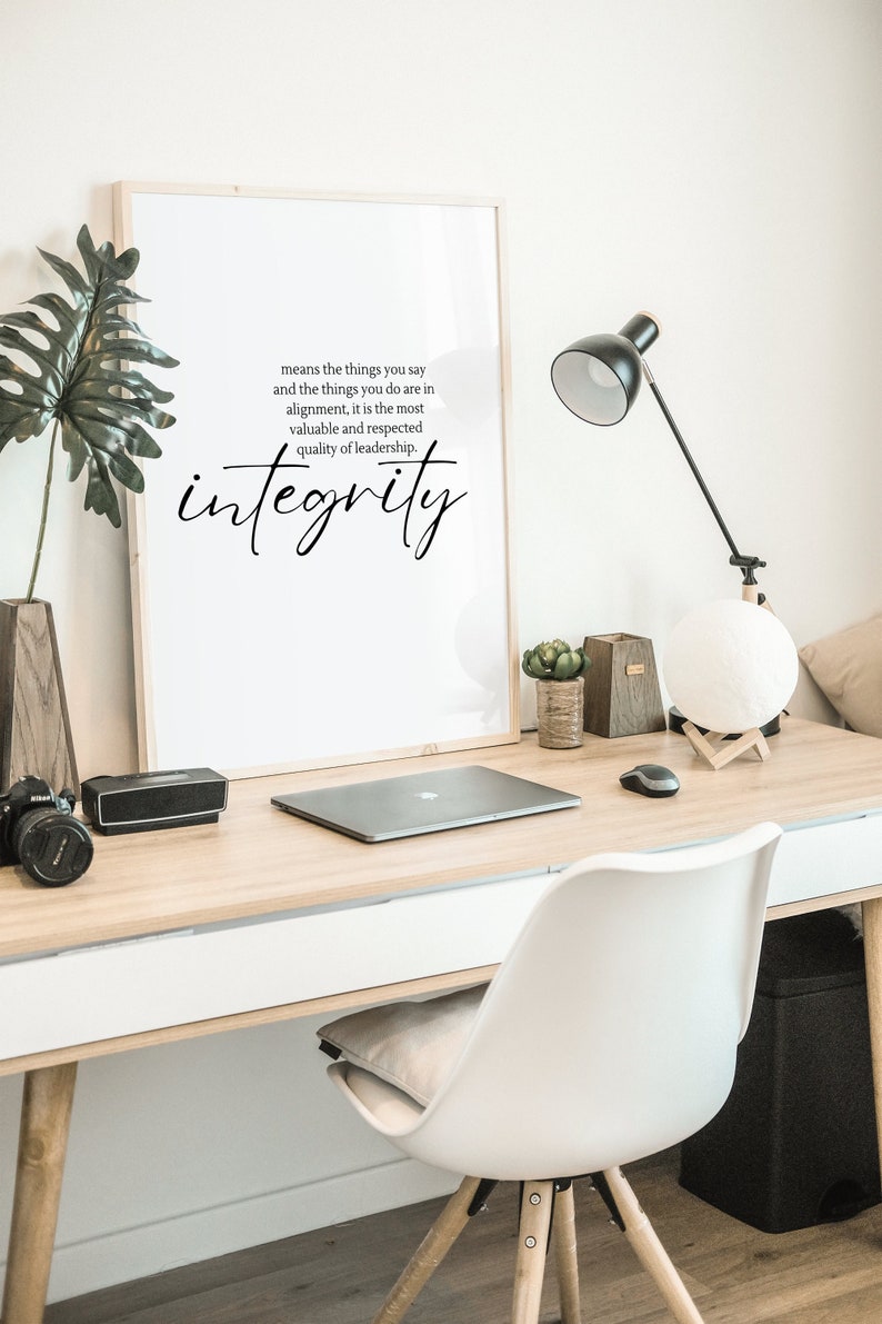 以下が含まれることがあります： 白い背景に黒い文字で「means the things you say and the things you do are in alignment, it is the most valuable and respected quality of leadership. integrity」と書かれた額装されたプリント。プリントは、ラップトップ、ランプ、鉢植えの植物のある机の上に置かれています。
