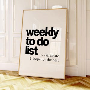 May include: A framed print with the words "weekly to do list" in bold black letters. The list includes "1- caffeinate" and "2- hope for the best". The print is in a light-coloured frame and leans against a white wall.