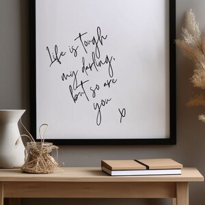 May include: Black framed print with a white background and black handwritten text that reads "Life is tough my darling, but so are you x". The print is sitting on a light brown wooden table with a white vase and a woven basket.
