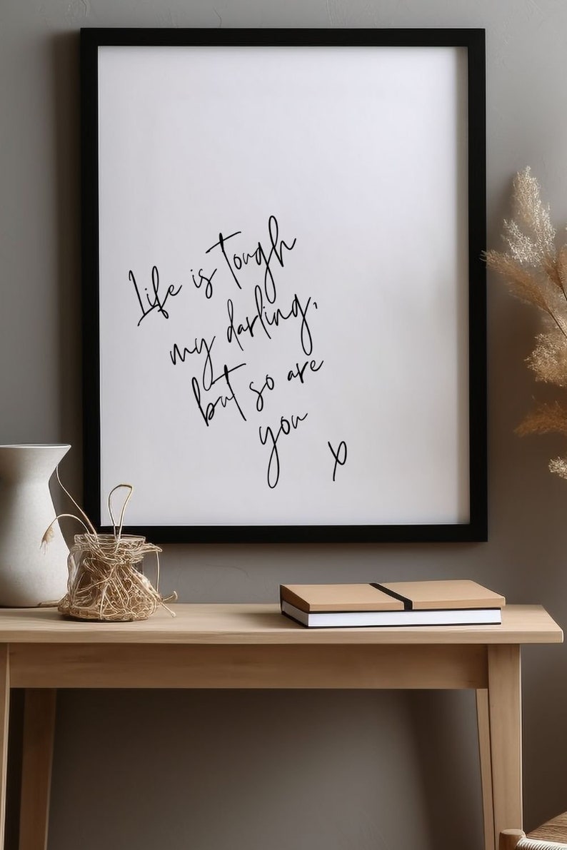 May include: Black framed print with a white background and black handwritten text that reads "Life is tough my darling, but so are you x". The print is sitting on a light brown wooden table with a white vase and a woven basket.