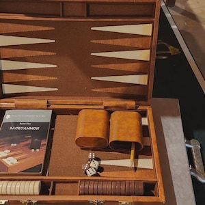 May include: A wooden backgammon set featuring a brown interior with cream and brown triangular points. The set includes two brown dice cups, a yellow pencil, white and brown checkers, dice, and an instruction booklet labeled Backgammon.