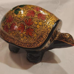May include: A turtle-shaped figurine with a glossy black base and a shell decorated with intricate gold, red, and green floral patterns. The head and legs are solid black with gold accents, and the entire surface features a smooth, reflective finish.