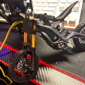 May include: A miniature black electric dirt bike model featuring gold-colored front suspension forks, black spoked wheels, and a detailed frame structure. The bike includes a small white logo on the side panel and a black seat, presented as a static display piece.