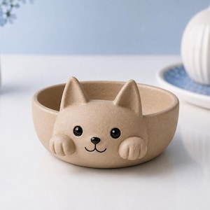 May include: A beige ceramic bowl featuring a sculpted cat face on the front rim. The cat design includes two pointed ears, two black eyes, a small black nose, a simple mouth line, and two front paws resting on the edge of the bowl's rim.