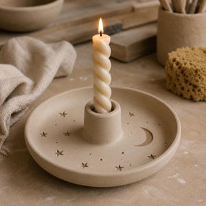 May include: A beige ceramic candle holder with a central pillar and a flat, circular base featuring engraved star and crescent moon motifs. A single, cream-colored, twisted taper candle is inserted into the center and is currently lit.