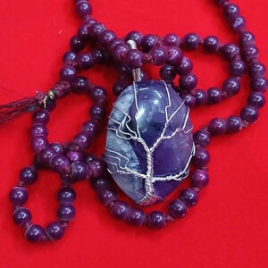Puede incluir: Un collar con un colgante de piedra ovalada pulida en tonos morado y blanco, envuelto en alambre de color plateado con forma de árbol. El colgante cuelga de una larga cadena de cuentas de madera redondas de color morado oscuro con una borla.