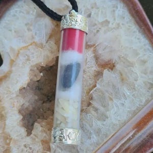 May include: A cylindrical pendant suspended from a black cord, featuring a clear glass tube encased in silver-toned metal caps with engraved star patterns. Inside the tube are stacked layers of red, white, black, and small off-white materials.