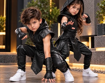 Può includere: Due bambini che indossano tuniche senza maniche con cappuccio, pantaloni e guanti senza dita coordinati in similpelle nera. Gli abiti presentano cinture strutturate e pannelli sovrapposti. Entrambi indossano scarpe da ginnastica bianche.