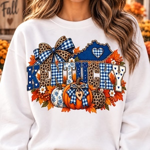 Peut inclure: Un sweat-shirt blanc à manches longues avec une impression graphique du mot KENTUCKY en motifs à carreaux bleus et léopard. Le design comprend le contour bleu de l'État, une grosse citrouille orange, un nœud à carreaux bleus et des feuilles d'automne.