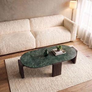 May include: An oval coffee table with a dark green marble top and four thick, dark brown wooden legs. On the table sits a small green glass vase and a stack of two books topped with a small green succulent plant in a white ceramic pot.