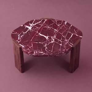 Puede incluir: Una mesa baja con un tablero hexagonal redondeado de mármol color burdeos intenso con vetas blancas prominentes. El tablero está sostenido por tres patas gruesas de madera marrón oscuro integradas en los bordes de la superficie.