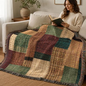 May include: A woven throw blanket featuring a patchwork design of rectangular blocks in shades of forest green, deep burgundy, and golden tan. The fabric has a textured, multi-tonal appearance with frayed edges along the sides and bottom.