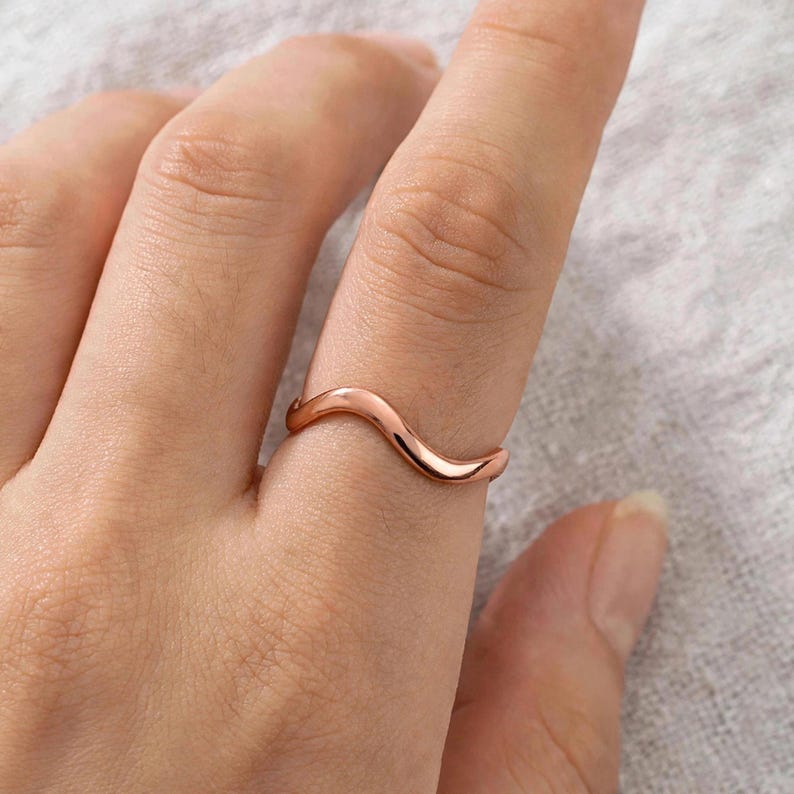 Handmade pure solid copper wave ring on index finger close-up natural skin texture detail light linen background minimalist lifestyle, thin organic wavy contour band polished curved ripple bypass design high mirror shine chevron stacking, modern boho ocean wave wishbone ring for women solid unplated copper everyday jewelry Jaipur artisan handmade pure copper ring gift for her beach lover