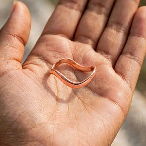 Handmade pure solid copper wave ring resting in open palm hand outdoor bright sunlight palm lines texture detail natural warm brown skin tone, organic thin wavy contour bypass band unplated copper high mirror polish ripple chevron stacking design minimalist boho ocean wave wishbone ring solid copper Jaipur artisan handmade everyday grounding jewelry gift for her wellness