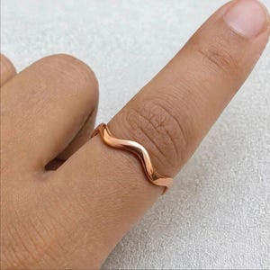 Handmade rose gold vermeil over 925 sterling silver organic wave ring on finger close-up neutral white textured linen studio light warm skin tone detail, minimalist thin wavy band polished ripple bypass contour high mirror shine chevron stacking design, modern boho ocean wave wishbone ring for women dainty surf inspired everyday inclusive jewelry Jaipur handmade rose gold wave ring gift