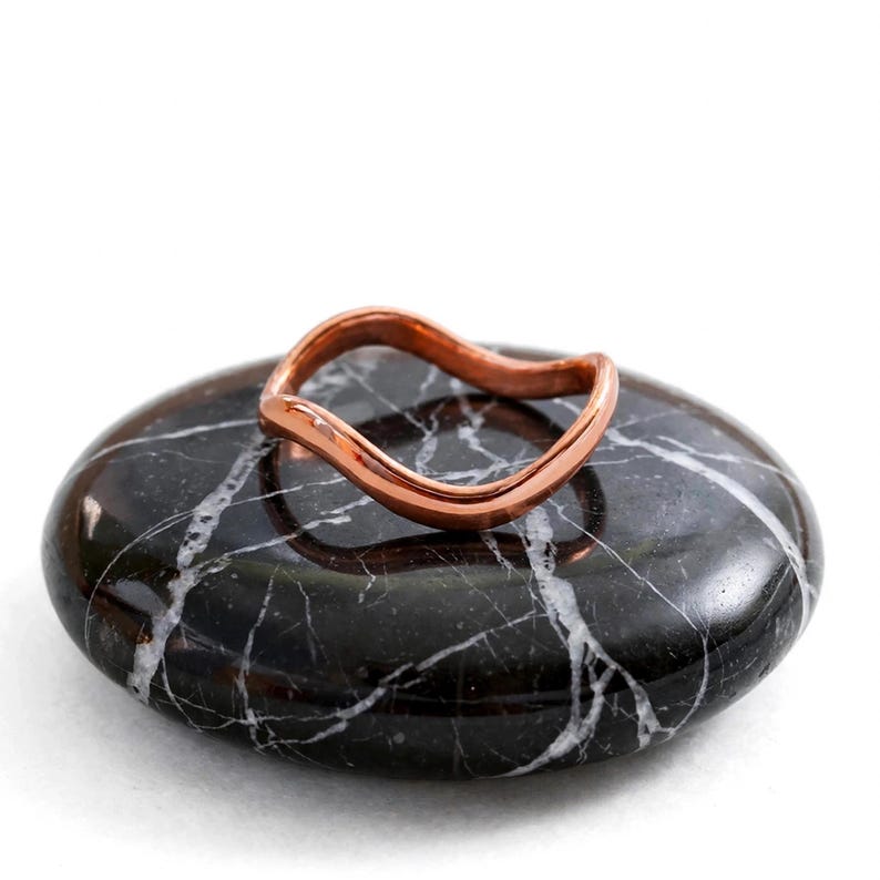Handmade pure solid copper wave ring resting on polished black marble pebble stone with white veining white background studio product shot, thin organic wavy contour band unplated natural copper high polish ripple chevron bypass stacking design reflection on stone, minimalist boho ocean wave wishbone ring solid copper Jaipur artisan pure copper ring zen spa display gift for her