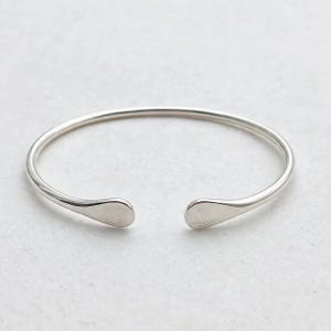 Puede incluir: Una pulsera abierta de tono plateado con un acabado liso y pulido. La pulsera presenta una banda circular que se estrecha en dos extremos redondeados y aplanados que se enfrentan con un pequeño espacio en el centro, formando un bucle.