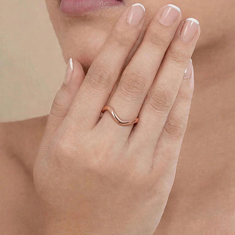 Handmade rose gold plated sterling silver wave ring on middle finger woman French manicure hand near chin lips beige background elegant close-up, minimalist thin wavy contour band polished ripple chevron bypass stacking design high mirror shine, modern boho ocean wave wishbone ring for women rose gold surf inspired everyday jewelry Jaipur handmade delicate rose gold ring gift for her