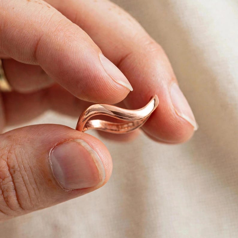 Handmade pure solid copper wave ring held between thumb forefinger bare natural short nails no polish extreme close-up beige linen blurred background, thin organic wavy contour bypass band unplated copper high mirror polish ripple chevron stacking curve shine detail, minimalist boho ocean wave wishbone ring solid copper everyday jewelry Jaipur artisan handmade unisex pure copper display gift