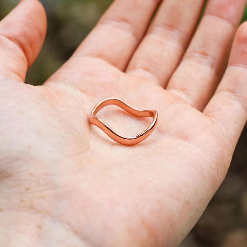 Handmade pure solid copper wave ring resting in open palm hand outdoor green blurred nature bokeh background natural daylight, thin organic wavy contour band unplated copper polished curved ripple high shine chevron bypass stacking, minimalist boho ocean wave wishbone ring solid copper everyday jewelry Jaipur artisan handmade pure copper palm display dainty surf gift for her
