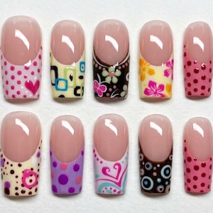 Puede incluir: Un juego de diez uñas postizas con estilo de manicura francesa, base color nude y diversos patrones coloridos en las puntas, incluyendo lunares rosas, cuadrados geométricos, estampados florales, círculos morados y motivos de corazones variados.