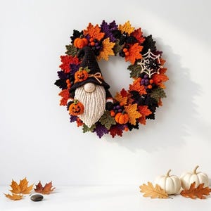 May include: A circular Halloween wreath featuring crocheted autumn leaves in orange, yellow, purple, and green. A central gnome figure wears a black hat with a small pumpkin, holding another pumpkin. Accents include black bats and a white spiderweb.