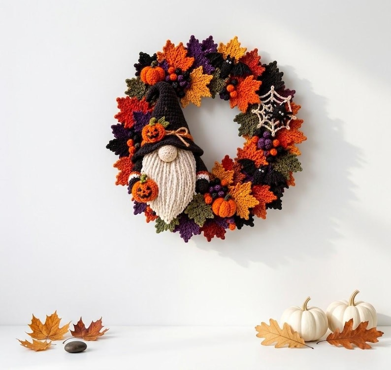 May include: A circular Halloween wreath featuring crocheted autumn leaves in orange, yellow, purple, and green. A central gnome figure wears a black hat with a small pumpkin, holding another pumpkin. Accents include black bats and a white spiderweb.