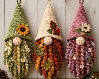 Pode incluir: Três gnomos de crochê pendurados com chapéus pontiagudos. O gnomo da esquerda tem um chapéu verde com um girassol, o central um chapéu creme com folhas de outono e bagas, e o da direita um chapéu malva com uma flor branca e um botão.