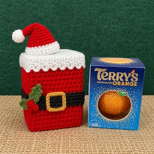 Può includere: Una copertura all'uncinetto rossa e bianca a forma di costume di Babbo Natale con cintura nera, fibbia dorata, foglie di agrifoglio verdi e bacche rosse, accanto a una scatola blu di Terry's Chocolate Orange con un'apertura circolare.