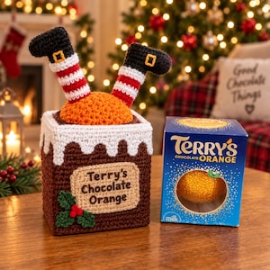 May include: A brown crocheted chimney cozy with white trim, green holly leaves, and red berries, featuring two legs in black boots and red-and-white striped socks sticking out the top, next to a blue Terry's Chocolate Orange box.