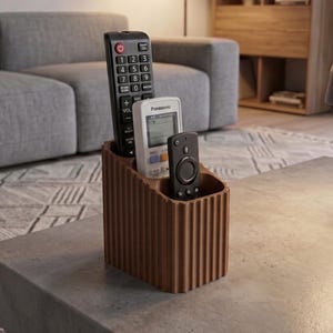 May include: A brown, rectangular remote control holder with a vertical ribbed texture on the exterior. The organizer features three distinct compartments, currently holding a black television remote, a white Panasonic remote, and a black streaming remote.