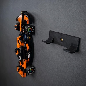 May include: A black and orange Formula 1 race car model featuring the text OKX and Chrome, mounted vertically on a wall. Beside the car is a black plastic wall mount bracket with two curved prongs and a central brass-colored screw insert.