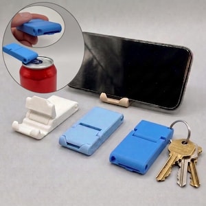 Könnte beinhalten: Ein Set aus faltbaren Handyhaltern aus Kunststoff in Weiß und Blau, wobei ein blauer Halter an einem Metallschlüsselring mit Schlüsseln befestigt ist. Ein schwarzes Smartphone liegt auf einem beigen Halter. Ein Inset zeigt einen blauen Halter beim Öffnen einer roten Dose.